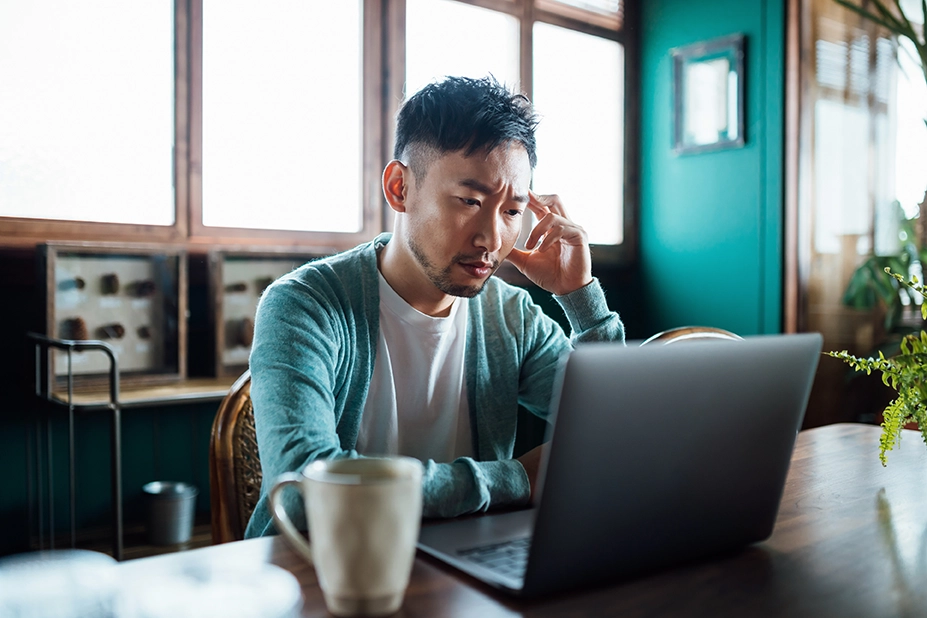Person working on a laptop at a table, resting head on one hand and looking concerned in a home or office setting with a coffee mug nearby